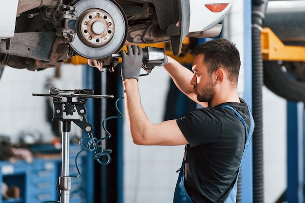 Met behulp van speciaal gereedschap man in werkuniform repareert witte auto binnenshuis conceptie van autoservice