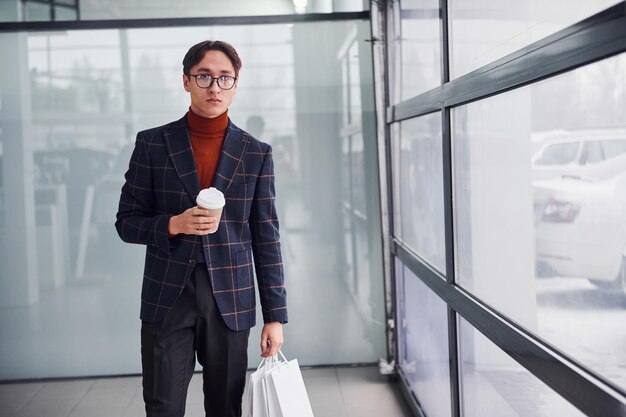 Met beker drinken en boodschappentassen. Jonge zakenman in luxe pak en formele kleding is binnenshuis op kantoor.