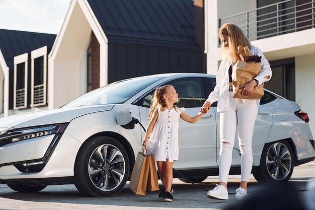 Met boodschappentassen Jonge vrouw met haar dochtertje is buiten met hun elektrische auto