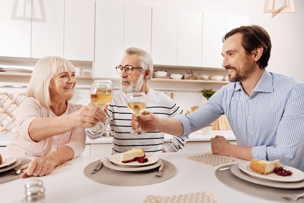 Met de beste wensen voor mijn ouders. Positieve gelukkig volwassen man aan het eten en genieten van vakantie met zijn bejaarde ouders terwijl hij glazen vol champagne opheft en glimlacht