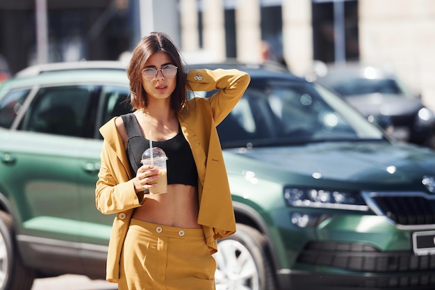 Met drankje in handen Jonge modieuze vrouw in bordeaux gekleurde jas overdag met haar auto