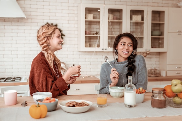 Met een brede glimlach. Glimlachend blond meisje ontbijten met haar glimlachende knappe vriend terwijl ze in de keuken zitten