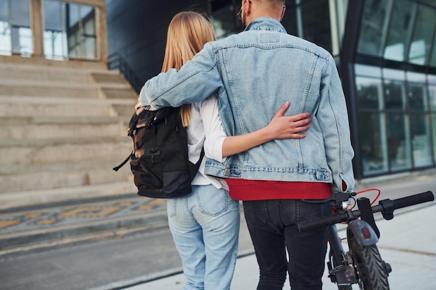 Met elektrische schooter Jonge stijlvolle man met vrouw in vrijetijdskleding buitenshuis in de buurt van zaken die samen bouwen Conceptie van vriendschap of relaties