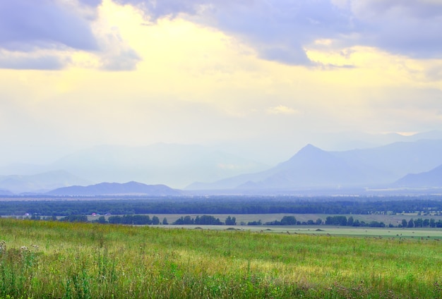 Met gras begroeide steppe tussen blauwe bergen in mist, bewolkte avondrood. Siberië, Rusland