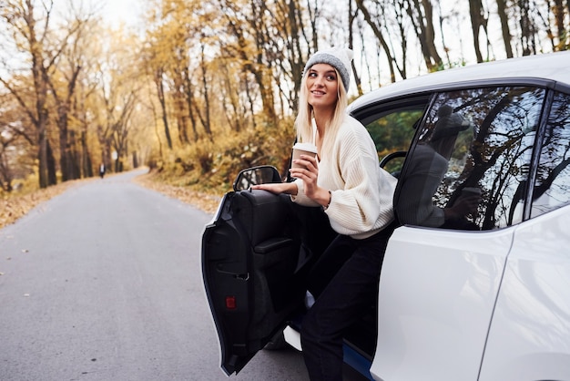 Met kopje drank in handen. meisje heeft herfstreis met de auto. moderne gloednieuwe auto in het bos.