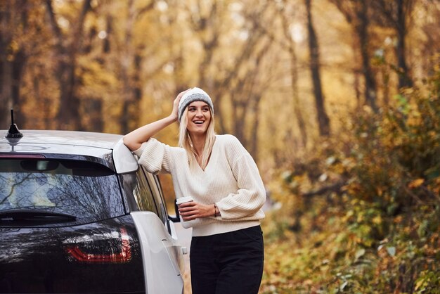 Met kopje drank in handen. Meisje heeft herfstreis met de auto. Moderne gloednieuwe auto in het bos.