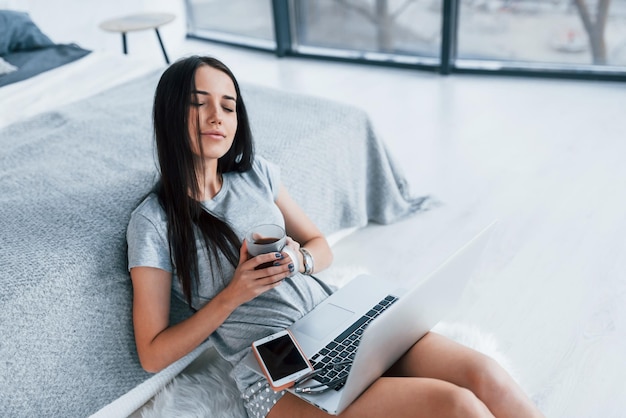 Met kopje thee en smartphone. Sexy jonge mooie vrouw liggend op bed met laptop.