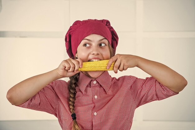 Met sterke tanden, gezond en biologisch voedsel, alleen vol vitamines, kind draagt chef-kok uniform en hoed tienermeisje bijt maïs verse rauwe groenten tijd om te eten gelukkig kind koken in de keuken