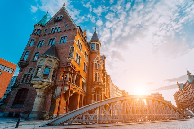 Metal arch bridge and old red bricks building in the speicherstadt warehouse district of hamburg hafencity with sunburst light during sunset golden hour and white clouds against blue sky above