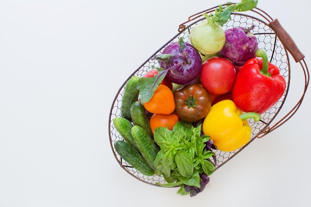 Photo metal basket with fresh vegetables on a white background fresh peppers tomatoes cucumbers basil leaves and kohlrabi in metal basket