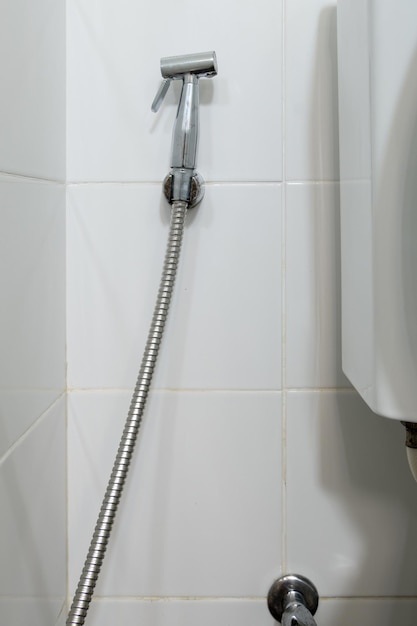 The metal bidet spray is hanging on the white tile wall near the flush toilet