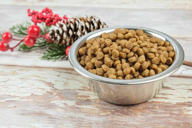 Metal bowl with dry pet food and Christmas decoration with red berries and pine cone on wooden boards. Christmas pet supplies concept, New year and Christmas celebration.