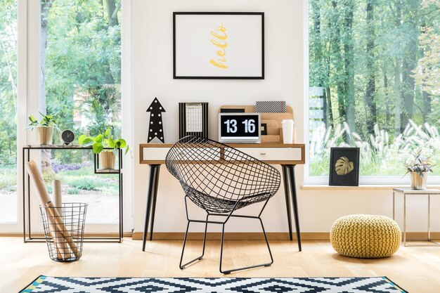 Metal chair at desk with laptop between yellow pouf and basket in workspace with poster and plants