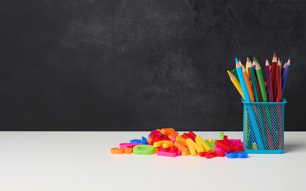 Metal glass with pens, pencils and felt-tip pens and scissors on the background of an empty black chalk board, white table, copy space