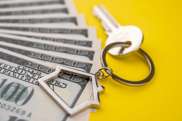 Metal key chain in the shape of a house with a metal key lying on the pack of hundred US dollars on a yellow background