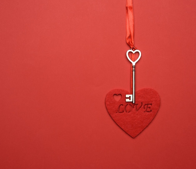 Metal key and red felt heart hang on red ribbon, red background