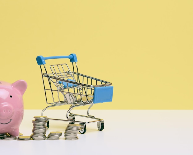 Photo metal miniature shopping cart with change and pink piggy bank on a white table yellow background concept of saving budget discounts