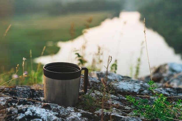 Metal mug on background of beautiful view on the morning calm river forest and hills Local tourism