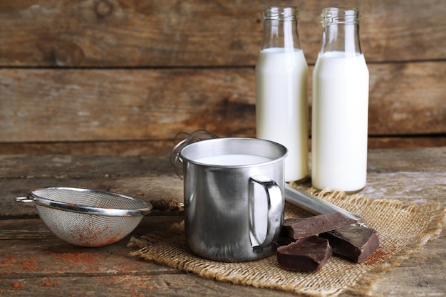 Metal mug and glass bottles of milk with chocolate chunks and strainer of cocoa on burlap cloth and rustic wooden planks background