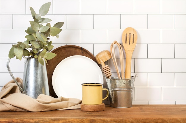 Metal mug and kitchen utensils on a wooden board in front of a subway tiles wall