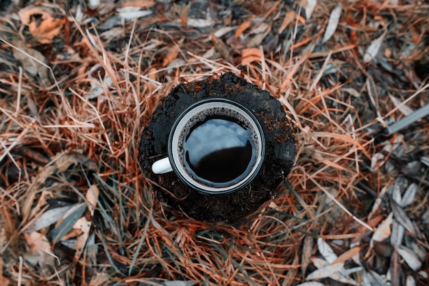 Metal mug with a hot drink stands on a tree stump in an autumn forest. Sky is reflected in the cup