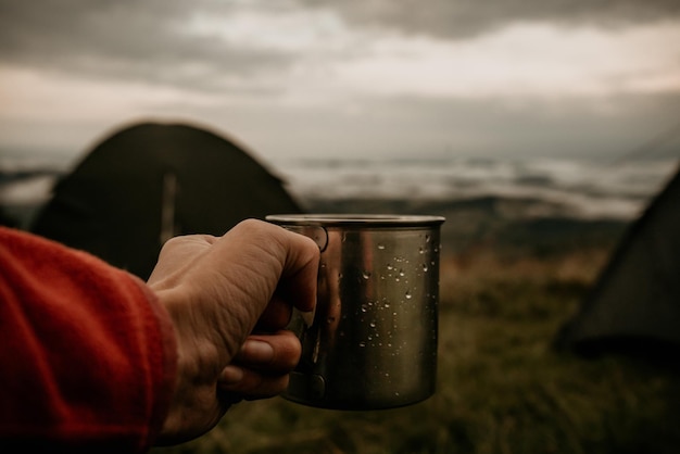 Metal mug with tea in hand of a tourist in tent camp in mountains under white clouds