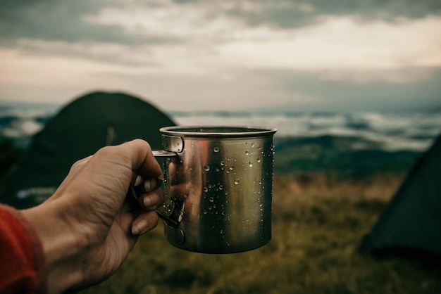 Metal mug with tea in hand of a tourist in tent camp in mountains under white clouds