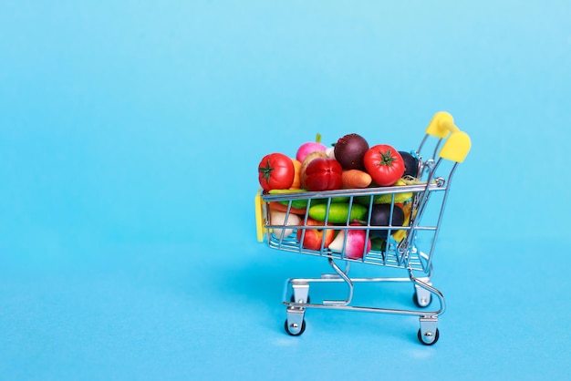 Metal shopping cart with fruits and vegetables on a blue background. Toy miniature shopping trolley