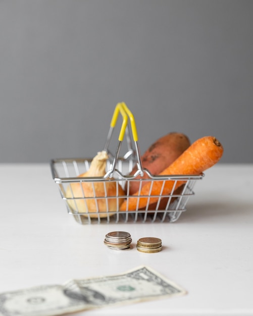 A metal supermarket food basket with vegetables and paper money and coins on a white table