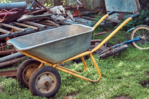 Metal wheel cart parked on grassy lawn near pile of shabby waste on summer day in countryside yard