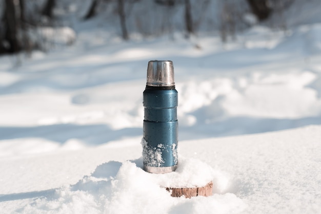 Metalen blauwe thermoskan staande op een besneeuwde boomstronk in een winter forest op een zonnige dag