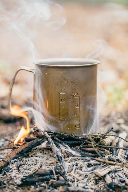Metalen campingmok met hete thee bij het vuur in het bos