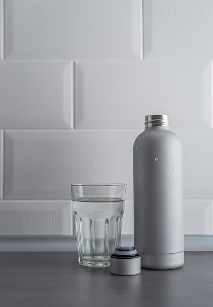 Metalen fles en glas water op grijs getextureerd aanrecht op een witte tegelmuur achtergrond