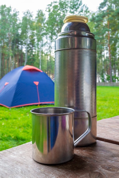 Metalen mok met thermoskan op houten tafel. blauwe toeristische tent op de achtergrond.