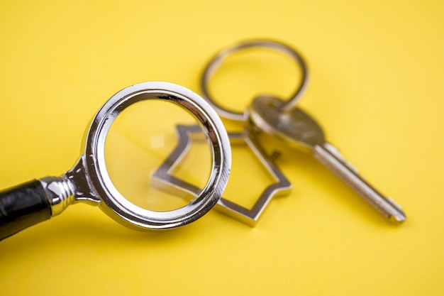 Metalen sleutelhanger in de vorm van een huis met een metalen sleutel onder een vergrootglas op een geel.