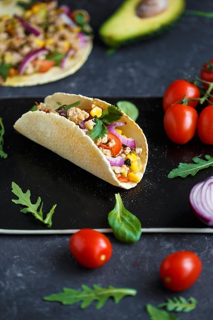 Mexicaanse taco's met varkensvlees, groenten, tomaten, avocado en kruiden op een zwarte stenen plaat op een donkere achtergrond met ingrediënten voor taco's