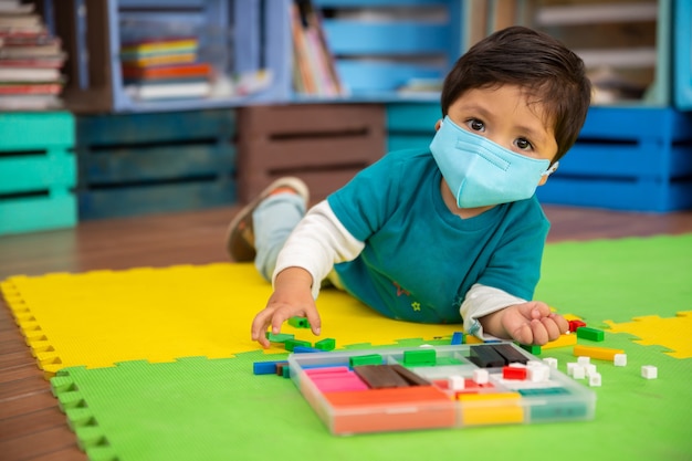 Mexican baby in school with mask playing with colored pieces on mat looking at the camera