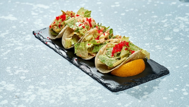 Блюдо мексиканской кухни - тако с лососем, листьями салата, белым соусом и икрой тобико на черной тарелке на синей поверхности