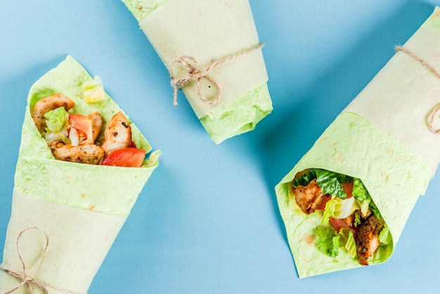 Photo mexican food. healthy eating. wrap sandwich: green lavash tortillas with spinach, fried chicken, fresh greens salad, tomatoes, yoghurt sauce. blue scene. copy space top view