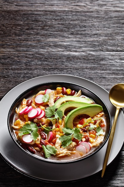 Mexican low carb chicken and tortilla soup with tomato, corn, red kidney beans, avocado, radish and lime, chile, fresh cilantro, on a black plate on a dark wooden background, top view, close-up