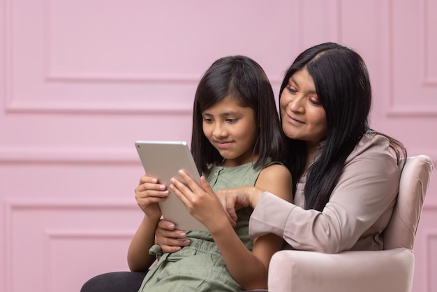Mexican mother and daughter using tablet isolated on pink background
