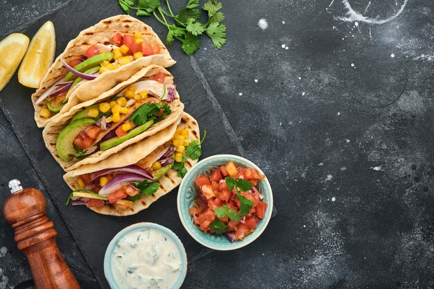 Mexican tacos with grilled chicken, avocado, corn kernels,\
tomato, onion, cilantro and salsa at black stone table. traditional\
mexican and latin american street food. top view.