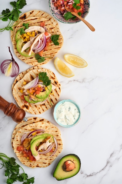 Mexican tacos with grilled chicken, avocado, corn kernels, tomato, onion, cilantro and salsa at white stone table. Traditional Mexican and Latin american street food. Top view.