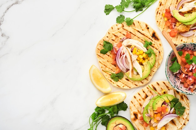 Mexican tacos with grilled chicken, avocado, corn kernels, tomato, onion, cilantro and salsa at white stone table. Traditional Mexican and Latin american street food. Top view.