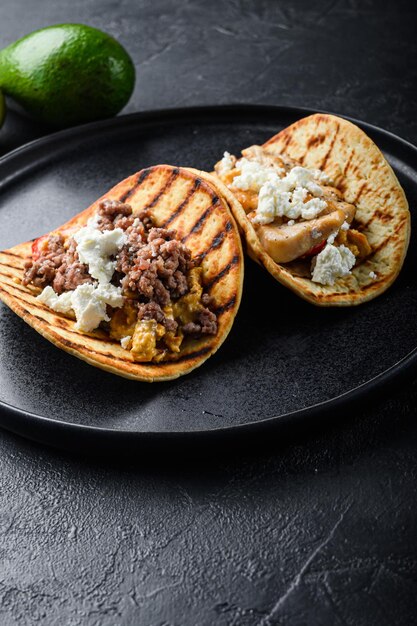 Mexican tacos with vegetables and beeaf and chicken meat on black round plate over textured black background side view selective focus vertical