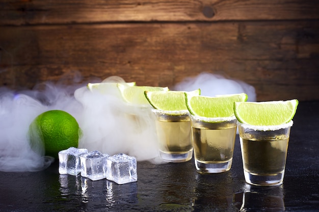 Mexican tequila gold in short glasses with salt, lime slices and ice on a wooden table. Smoke.