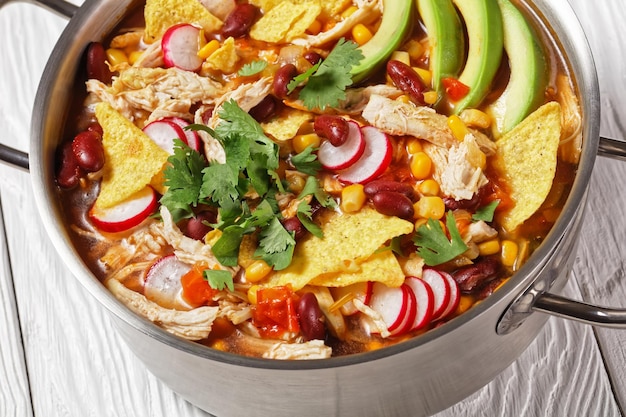 Mexican tortilla soup with pulled chicken, tomato, corn, red\
kidney beans, avocado, radish and lime, chile, cilantro, served on\
a metal pot on a white wooden background, top view, close-up