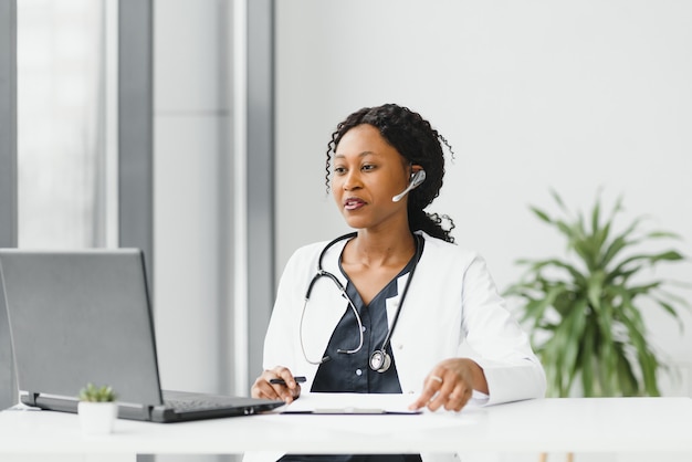 Mhappy lachende Afro-Amerikaanse vrouwelijke arts of verpleegkundige met hoofdtelefoon en laptop met conferentie of video-oproep in het ziekenhuis