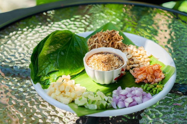 Miang kham закуска из королевских листьев - это традиционная закуска юго-восточной азии из таиланда и лаоса. она находится на белом блюде на открытом столе с солнечным светом.