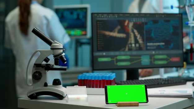 Microbiologist bringing blood samples in modern lab putting them near smartphone working with green chroma key screen placed on desk. Team of biotechnology scientists developing drugs in background